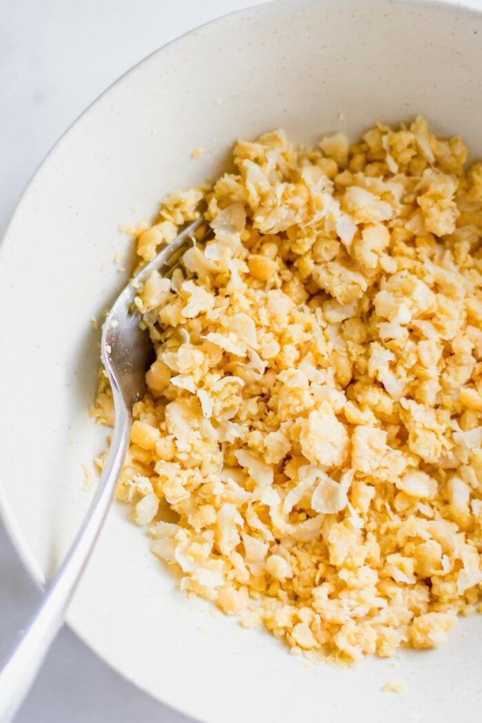 Mashed chickpeas in a large white mixing bowl with a fork resting in the bowl.