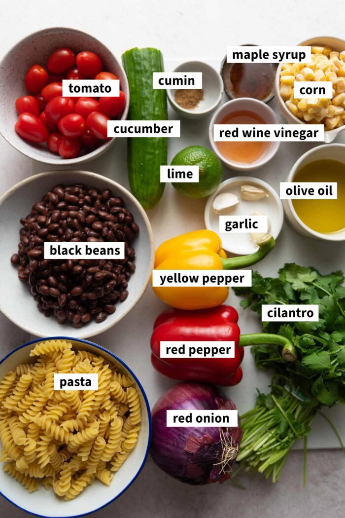 All the ingredients needed for a black bean pasta corn salad with tomato and cucumber, each labelled with text.