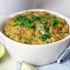 A bowl of vegan curried lentils with quinoa topped with chopped cilantro with a sliced lime beside the bowl and a pot of lentils in the background.