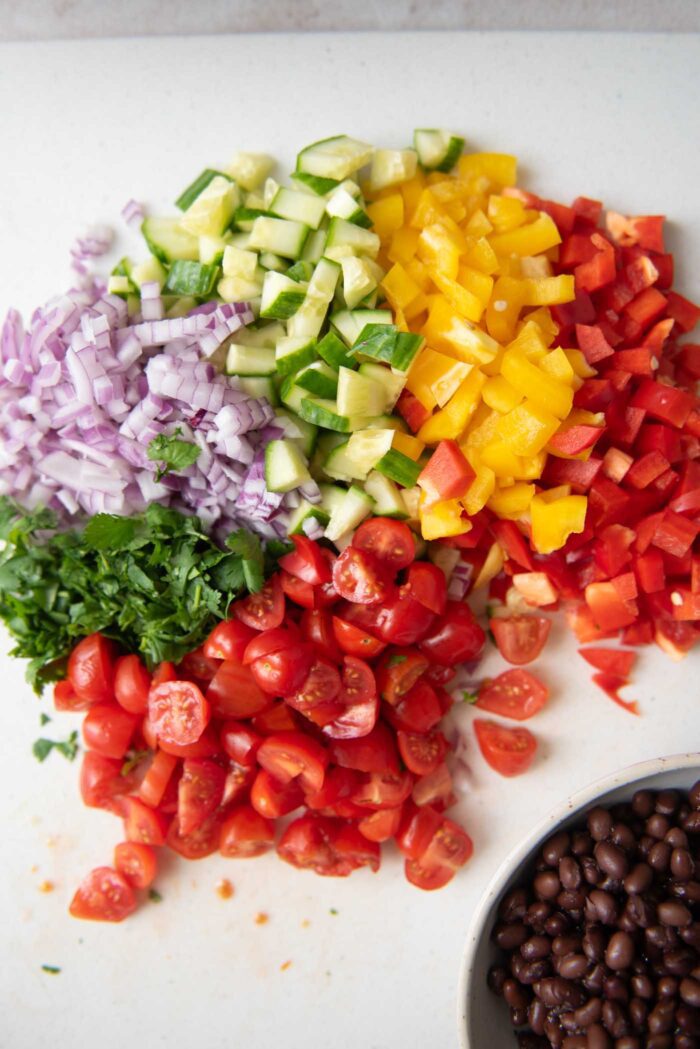 Chopped bell pepper, red onion, cilantro, tomatoes, cucumber on a cutting board.