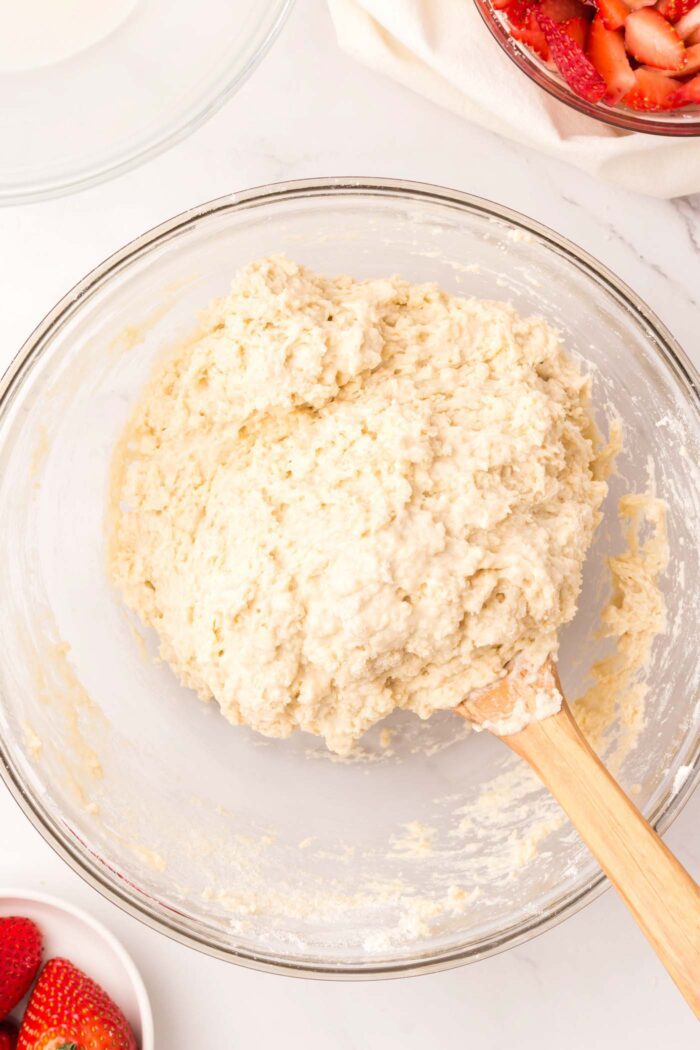 A thick biscuit batter in a glass mixing bowl with a wooden spoon.