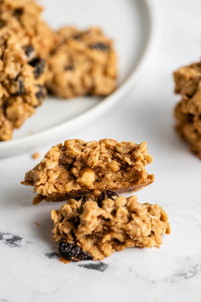 Small oatmeal cookie broken in half to show the inside texture. More cookies appear in the background on a plate.