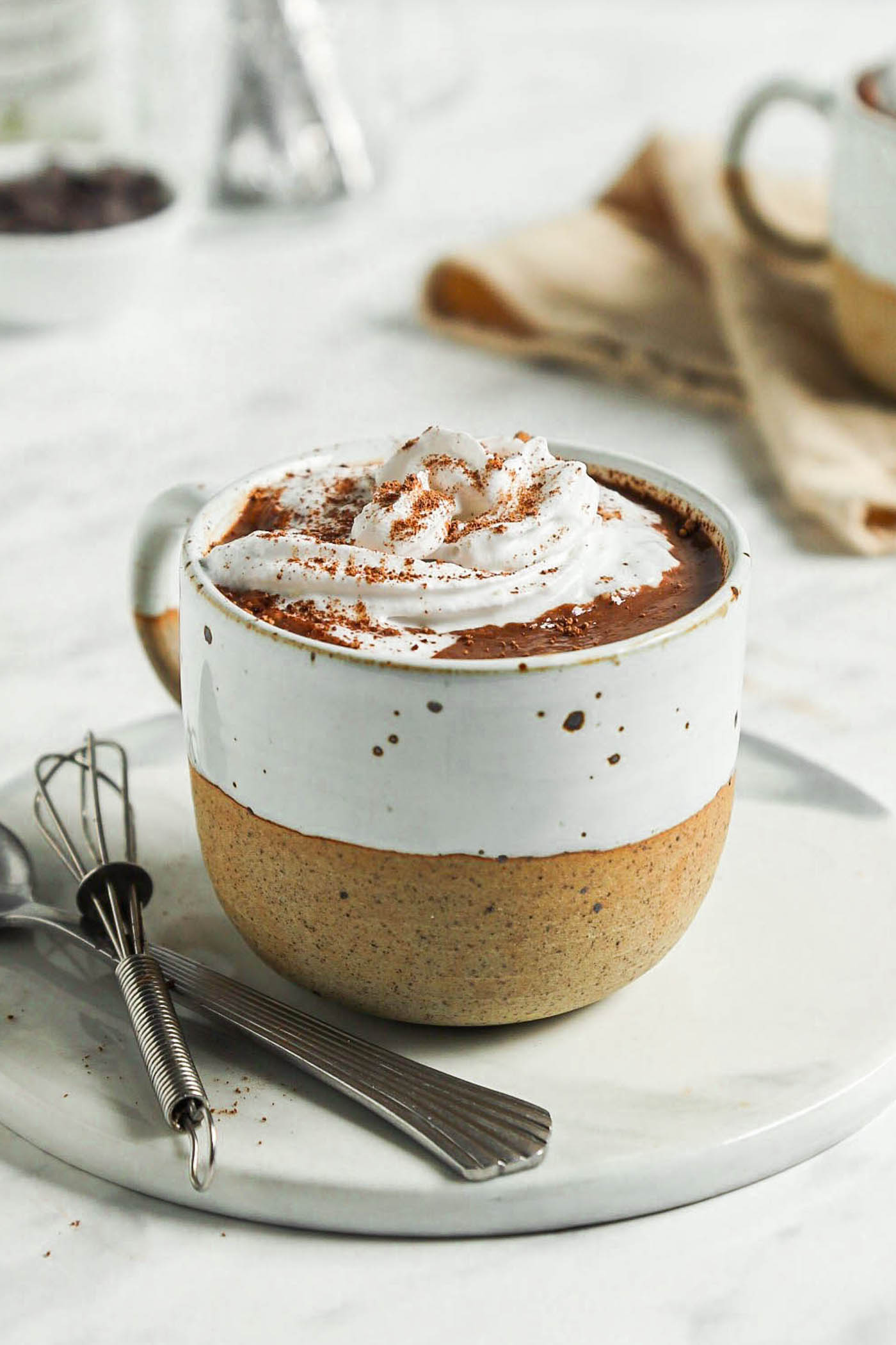 Close up of a mug of hot chocolate topped with whipped cream on a small plate with a spoon and a small whisk.