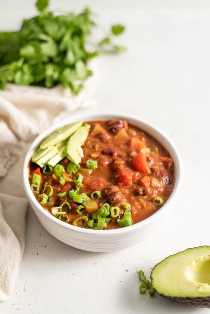 A bowl of vegan chili topped with avocado and green onion with half an avocado sitting beside it.