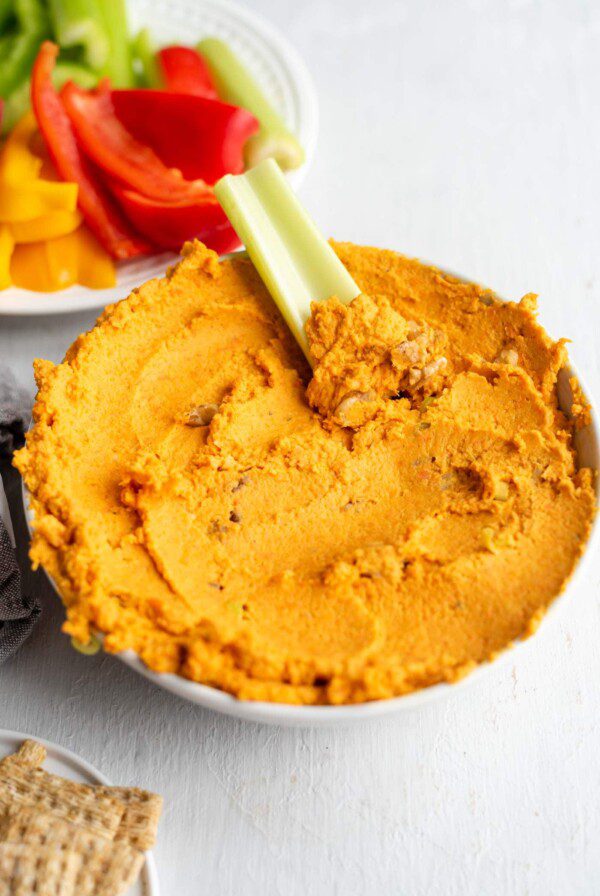 A bowl of roasted carrot hummus with a piece of celery dipped in it.