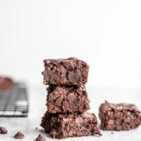 Stack of 3 vegan oat flour brownies on a grey surface with some chocolate chips scattered around.