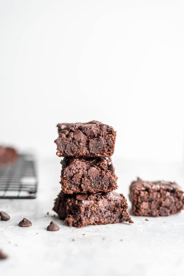 Stack of 3 vegan oat flour brownies on a grey surface with some chocolate chips scattered around.