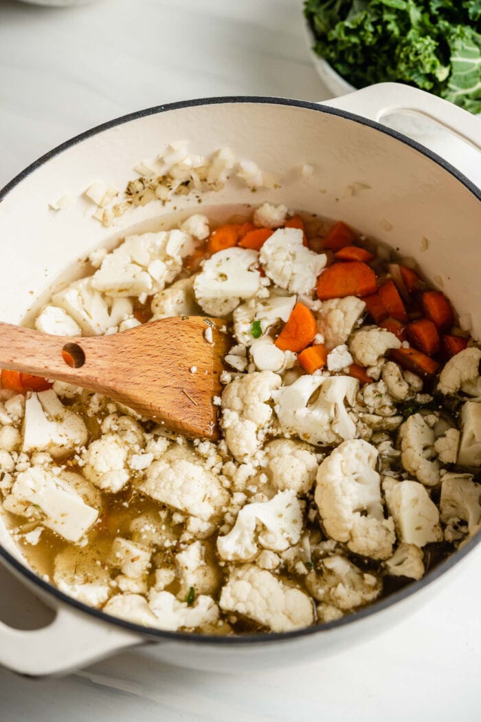 Chopped cauliflower and carrot cooking in a large soup pot with a wooden spoon.