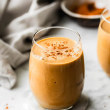 A creamy sweet potato smoothie in a glass sprinkled with cinnamon with a dish cloth laid out behind it.