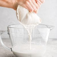 Straining the blended oat milk through a nut milk bag into a glass measuring cup.