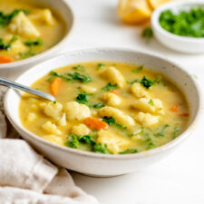 Bowl of kale and cauliflower soup with a spoon in it. Slices of lemon, dish of parsley and another bowl of soup in background.