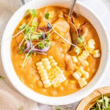 Overhead view of a handled bowl summer corn chowder soup topped with micro-greens and kernels of fresh corn. A spoon rests in the bowl and two additional bowls of soup are just out of frame.