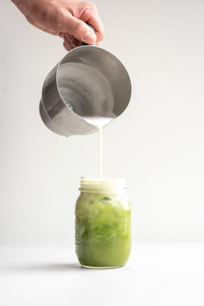 A hand pouring frothed almond milk into an iced matcha.