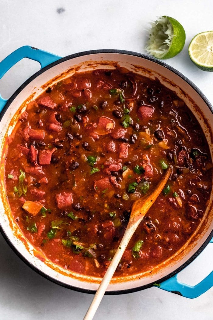 Chunky vegan tortilla soup in a soup pot with a wooden spoon and a lime on the side.