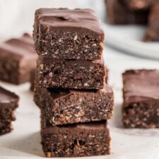 Stack of 4 no-bake hemp see brownies topped with chocolate frosting with more brownies scattered in the background.