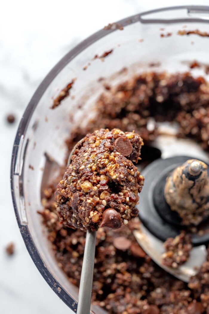 Raw chocolate brownie dough on a small spoon held over a food processor container of brownie batter..