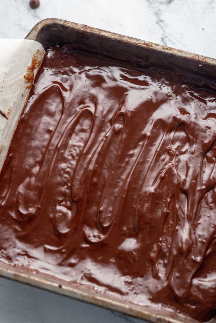 A thick layer of vegan chocolate ganache spread over raw hemp seed brownies in a square baking pan lined with parchment paper.