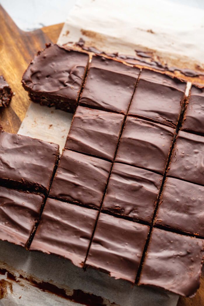 Raw vegan brownies topped with a layer of chocolate ganache cut into 16 squares on a round cutting board.