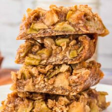 Stack of 4 apple crumble bars on a plate. The bars have an oatmeal crust and crumble topping with a layer of apple pie filling in between.