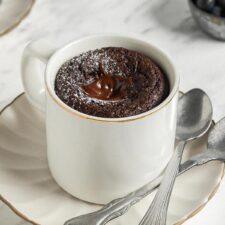 Chocolate mug cake on a small plate with 2 spoons resting on plate.