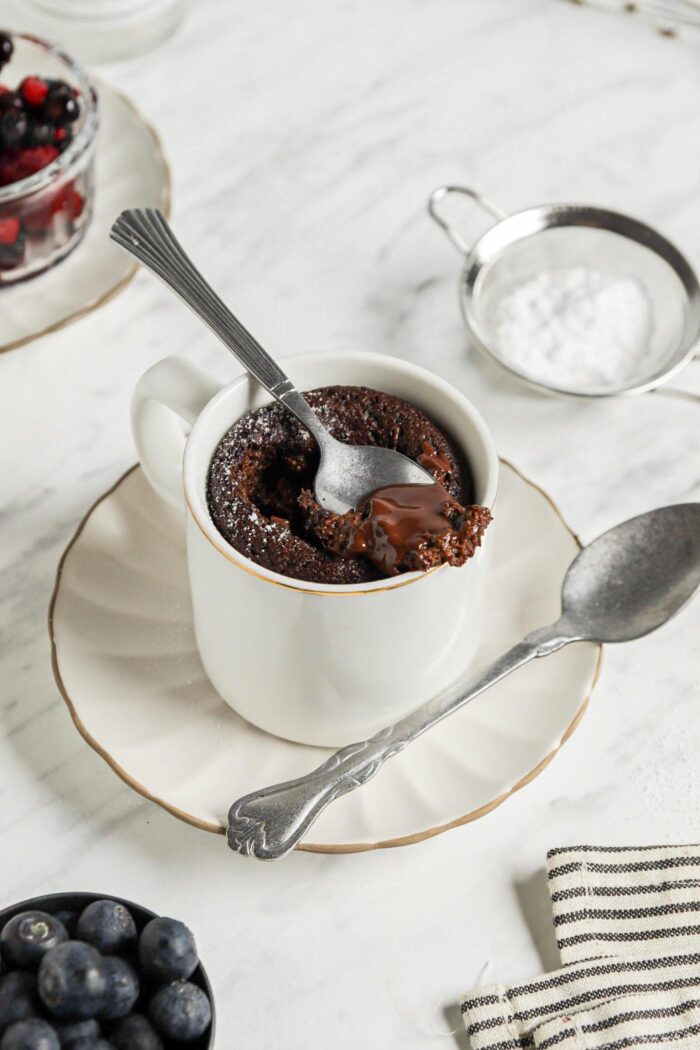 Spoon taking a spoonful of chocolate mug cake from a mug sitting on a small plate.