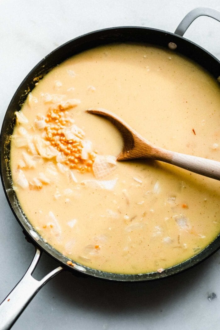 Coconut milk being stirred with lentils and onions in a skillet with a spoon.