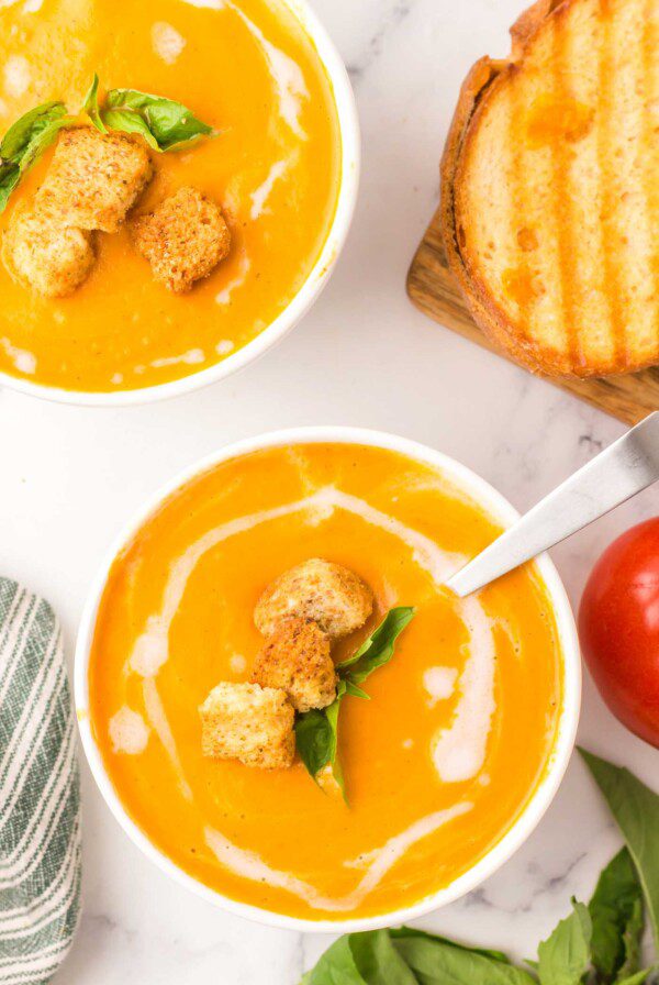 Two bowls of creamy vegan tomato soup topped with a drizzle of coconut oil and two croutons with a few slices of toasted bread beside them.