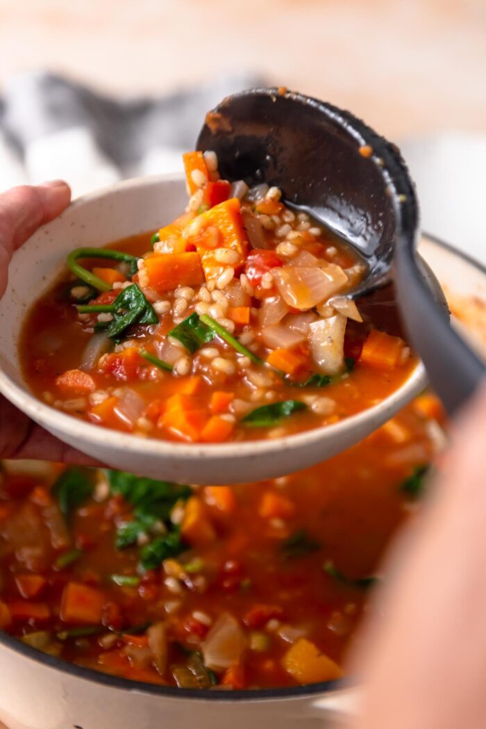 Scooping tomato vegetable barley soup from a pot into a serving bowl.