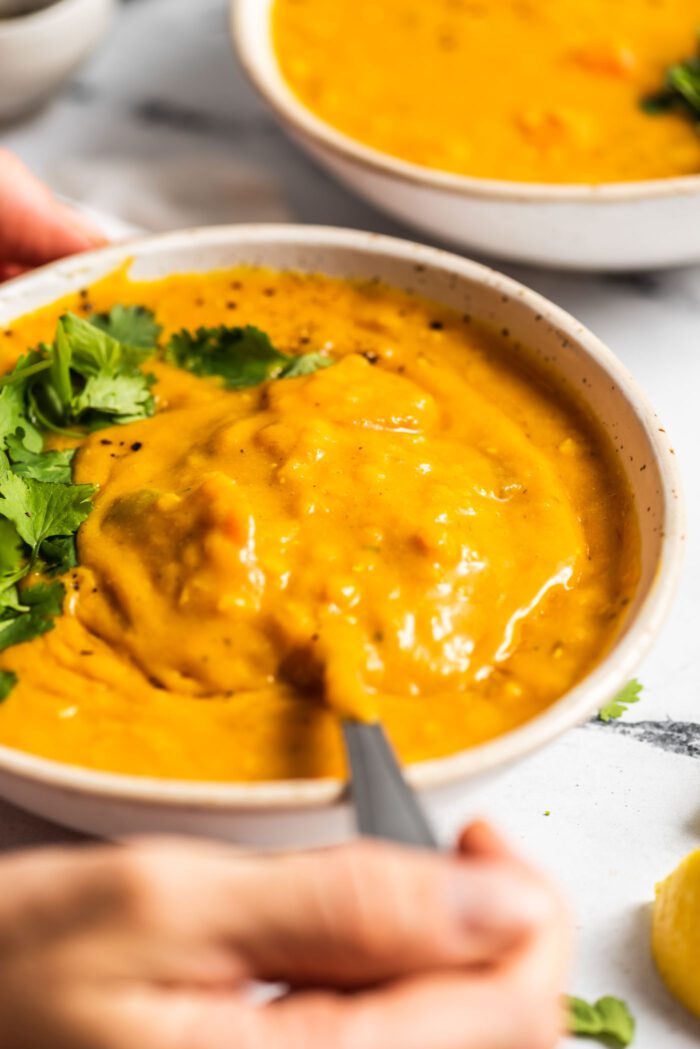 Hand using a spoon to lift a spoonful of lemon lentil soup topped with fresh cilantro from a bowl. Another bow of soup is in the background.