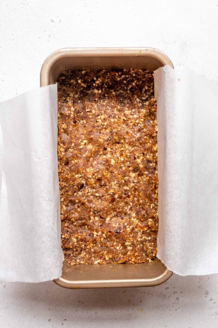 A raw walnut date crust pressed into a loaf pan lined with parchment paper.