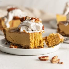 Slice of pumpkin cheesecake topped with whipped cream on a small plate. Bite taken out of slice of cake with a fork.