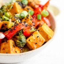 Pair of chopsticks rests on a bowl of stir fried vegetables and tofu in orange sauce.