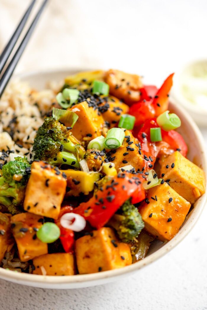 Close up of a bowl of orange tofu and vegetable stir fry topped with black sesame seeds.