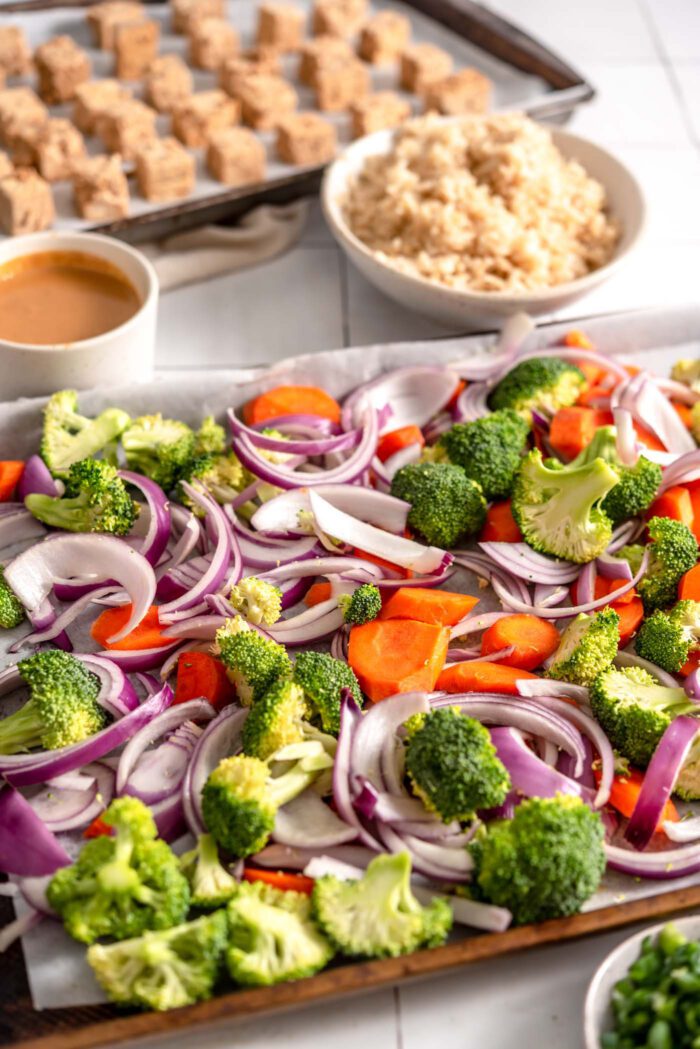 Chopped broccoli, onion and carrot on a baking pan. In the background, a pan with cubed tofu on it is visible.