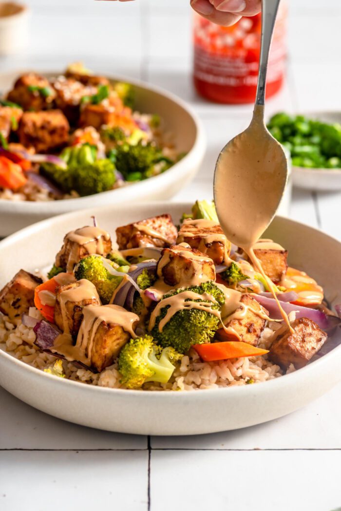 Spoon drizzling peanut sauce over a bowl of rice, vegetables and tofu.
