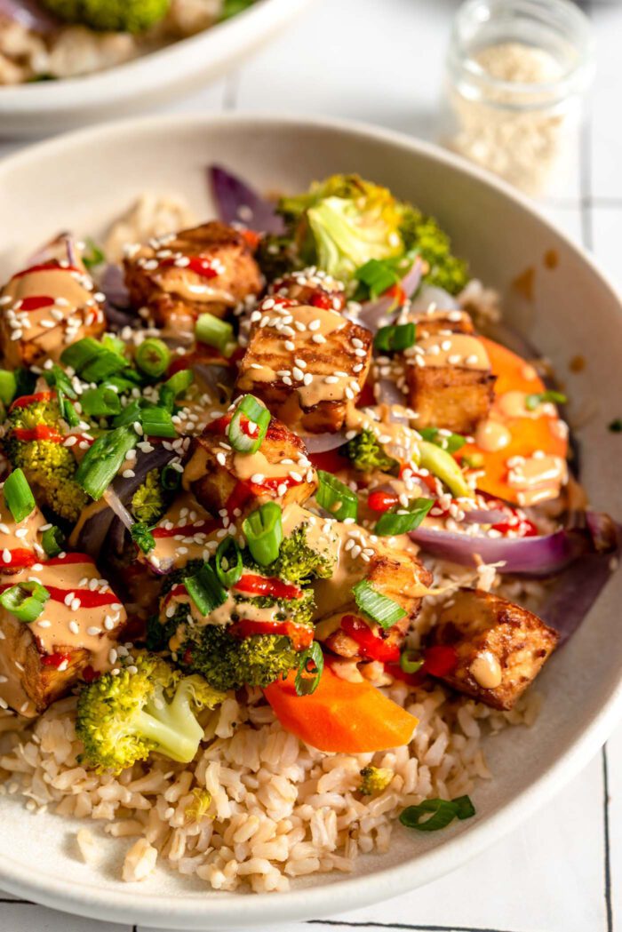 A colourful bowl with tofu, broccoli, carrot, onion and rice, topped with creamy peanut sauce, green onion, sesame seeds and hot sauce.