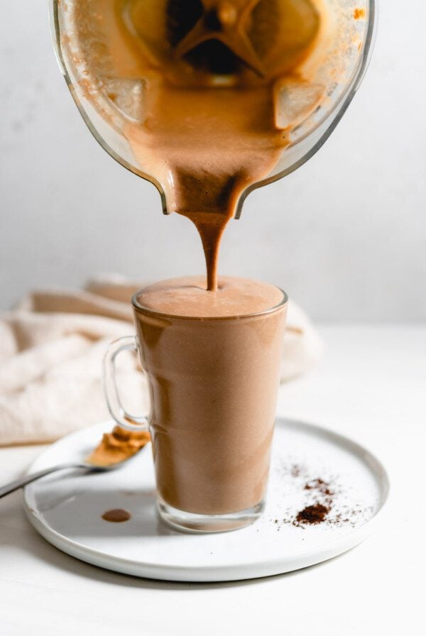 A blender pouring a chocolate smoothie into a glass mug sitting on a plate.