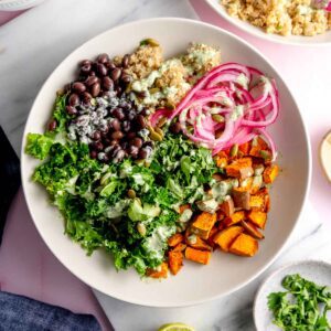 Bowl with roasted sweet potato, kale, black beans, quinoa, thinly sliced red onion and a creamy green sauce on top.