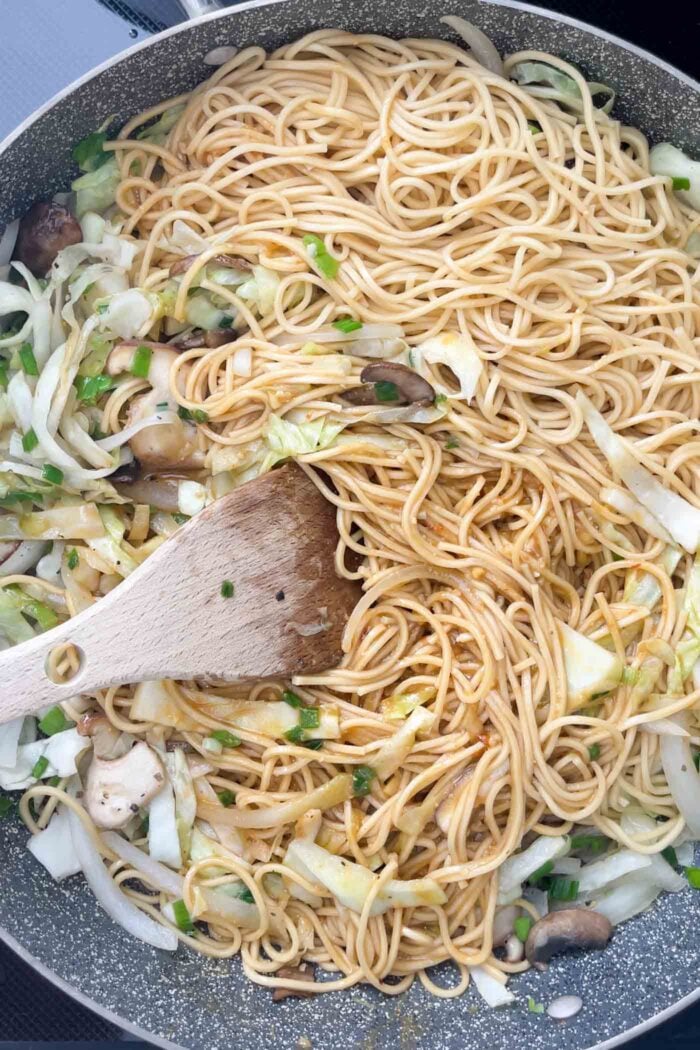 Noodles and sauce with cabbage and mushroom cooking in a large skillet with a wooden spoon.