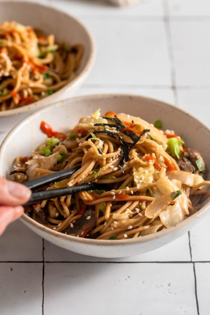 Hand using a set of chopsticks in a small bowl of miso noodles with cabbage and mushrooms.