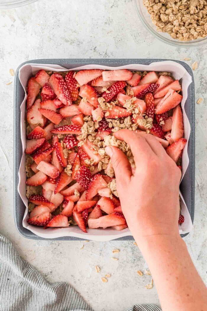 A hand sprinkling an oatmeal crumble mixture over a diced strawberries in a square baking pan lined with parchment paper.