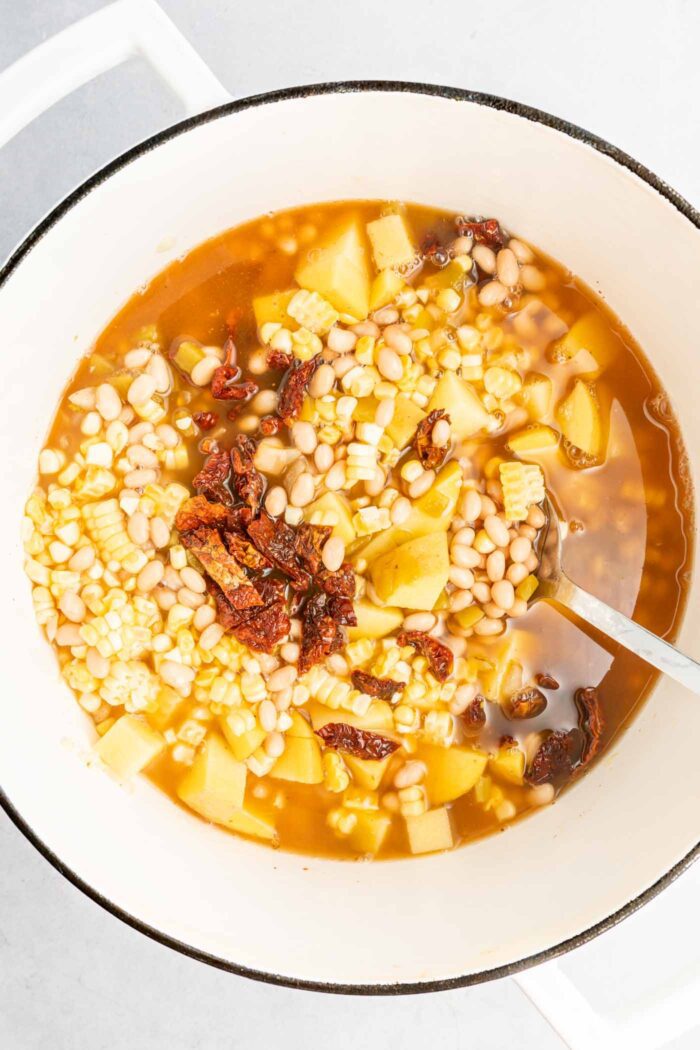 A large soup pot of corn, sun dried tomatoes, potato, white beans and broth. A spoon rests in the pot.