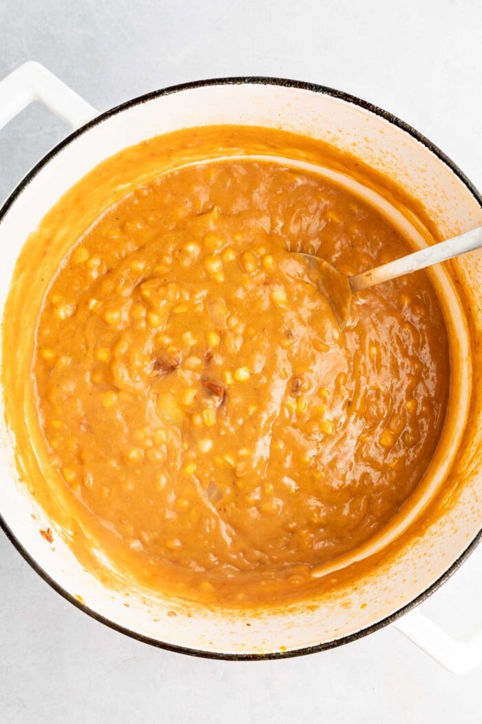 A large pot of creamy summer corn and potato chowder with corn kernels in it. A spoon rests in the pot.