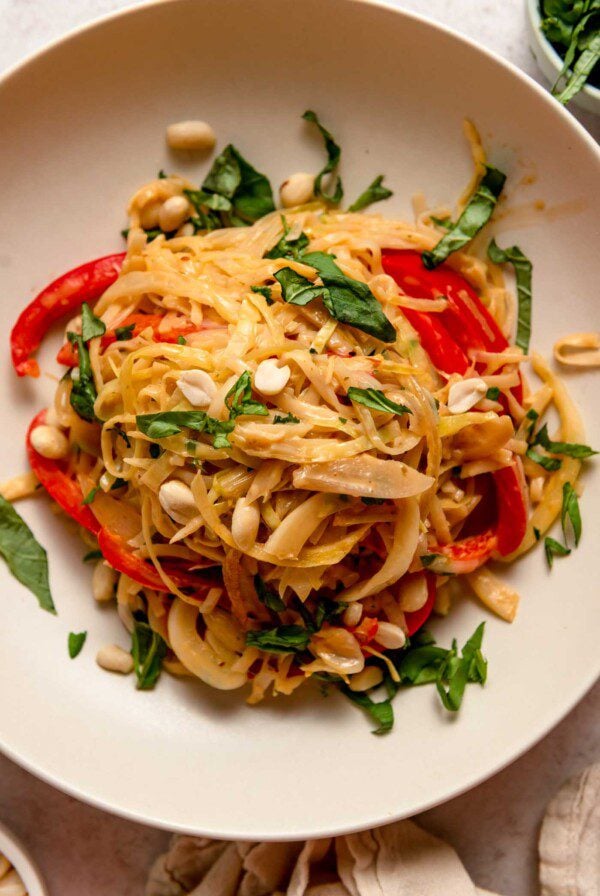 Overhead view of a bowl of red curry noodles with sliced red bell peppers, cilantro, peanuts and onion.