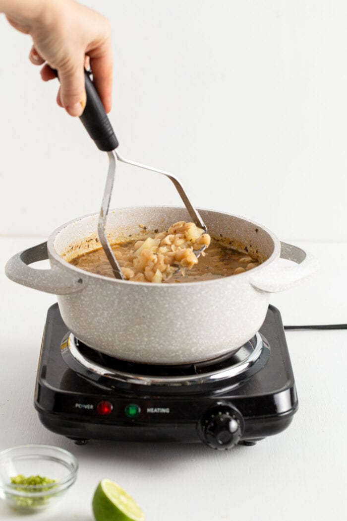 Mashing a pot of potato and beans with a potato masher in a pot.